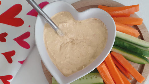 Homamade humus in a white heart ramekin with raw carrots and cucumber