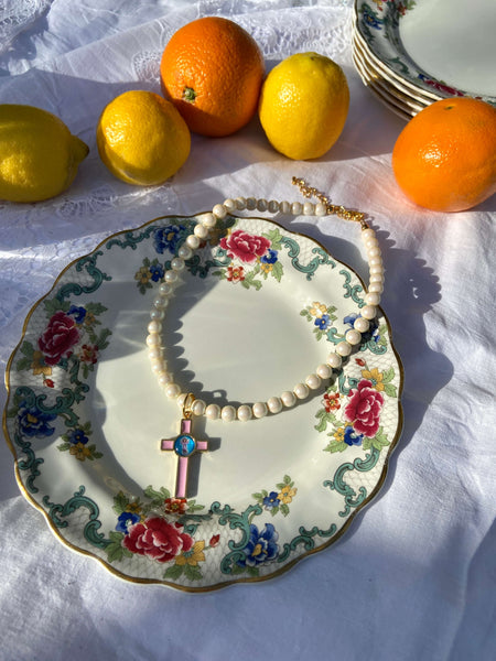 A round pearl necklace in white with a pink cross pendant on a white picnic cloth surrounded by oranges and lemons 