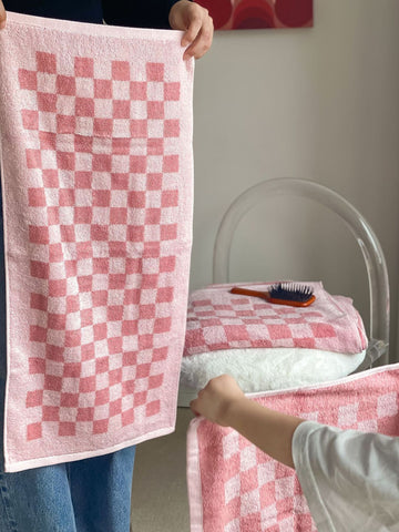 A woman and child holding pink, checkerboard-printed towels. A chair sits in the background, holding another pink, checkerboard-printed towel on its seat with a comb on top of the towel