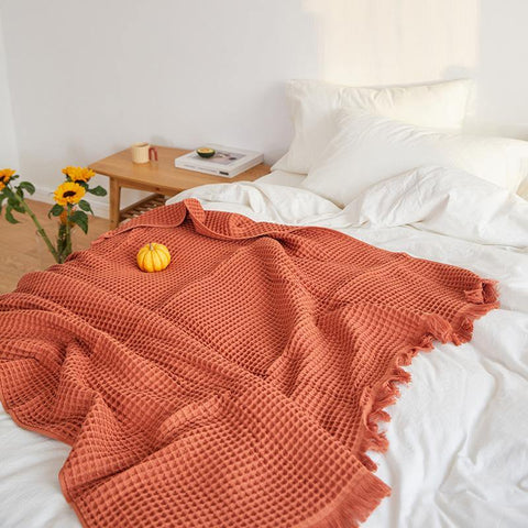 A tomato-coloured Fringed Waffle Throw draped over a bed with white bedding sheets. There is a small pumpkin decoration piece on top of the blanket throw.