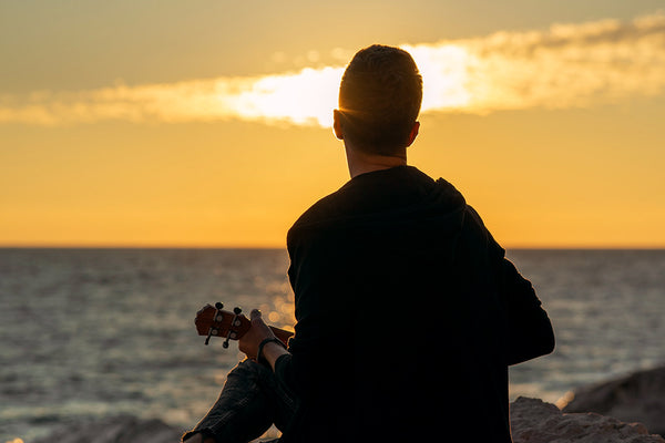 Mann mit Ukulele vor Sonnenuntergang am Meer