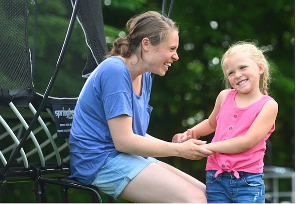 A mother holding her daughter's hand while sitting on a trampoline ladder, both smiling.