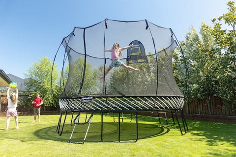A girl jumping on a Springfree Jumbo Round Trampoline while two kids play ball outside of it.