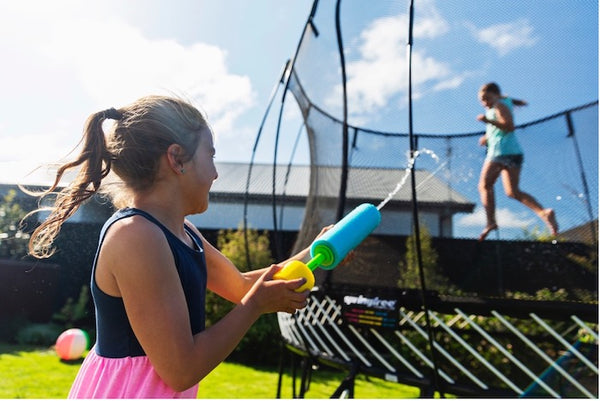 A little girl spraying a water gun at another girl jumping on a trampoline.