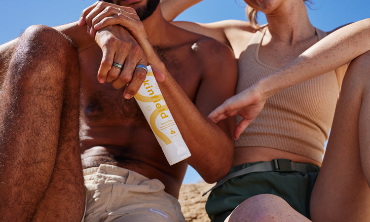 Close up image of two torsos, with bare arms sitting in the sun, holding a white and yellow tube of Kinfield Cloud Cover Body SPF 35