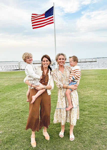 Pepa holding her son, Matthew, and Jenifer holding her son, Hudson, standing outside on the waterfront in front of an American flag.