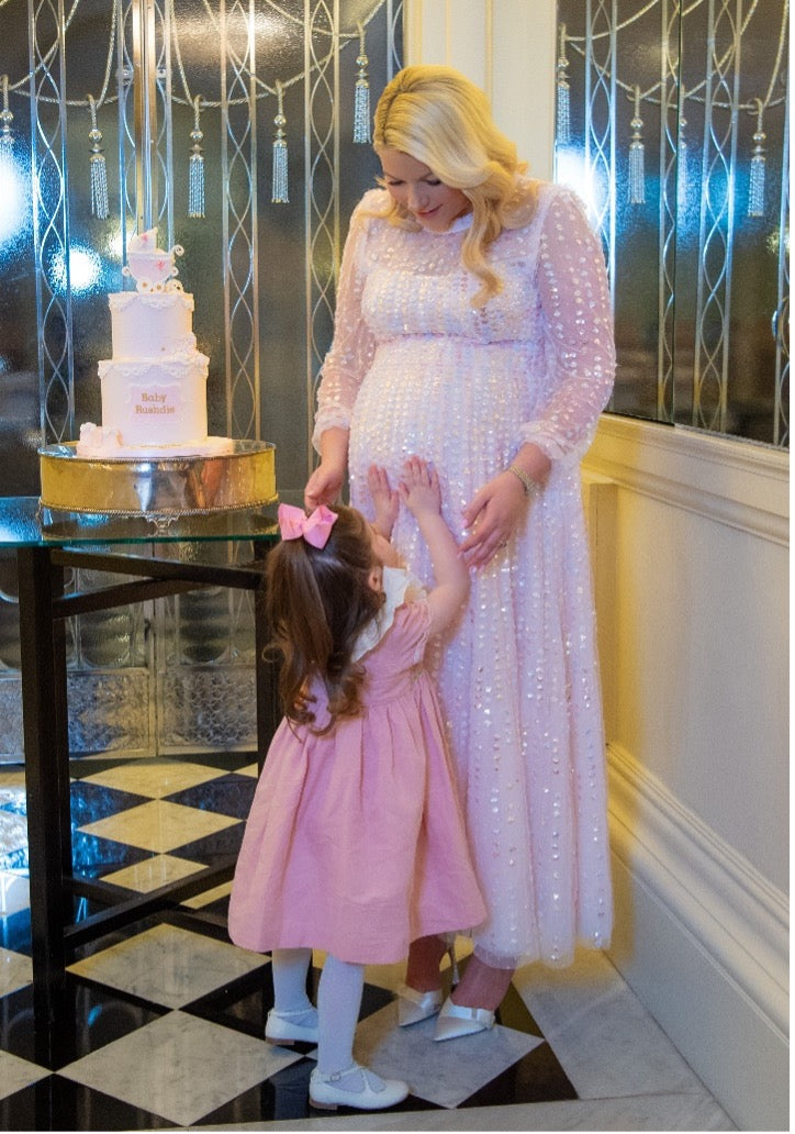 Natalie Rushdie standing in a white dress in the corner of a room with black and white check tiles, in front of a table with a white tiered cake. Her daughter looks up at Natalie, touching her pregnant belly.