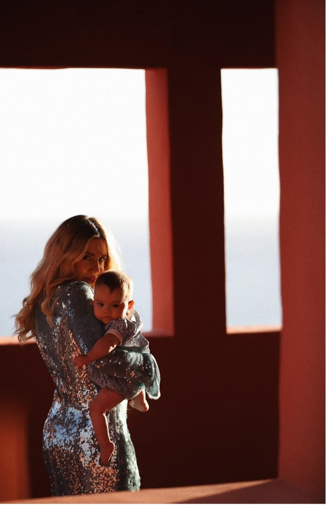 Sara-Louise is standing in front of a window, wearing a silver sequinned dress and holding her baby in her arms.