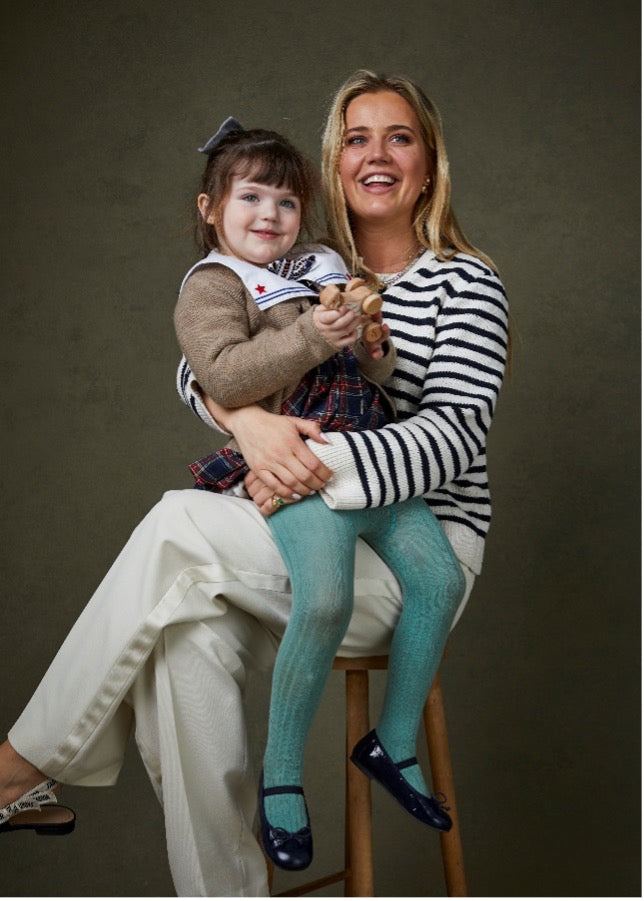 Porträtbild von Tabitha, die mit gekreuzten Beinen auf einem Hocker sitzt und ihre kleine Tochter auf ihrem Schoß sitzt.