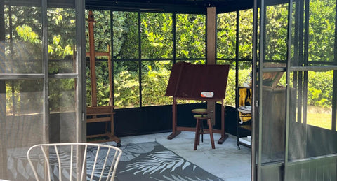 A bright sunroom, enclosed by screens and windows, is set up as an art studio.