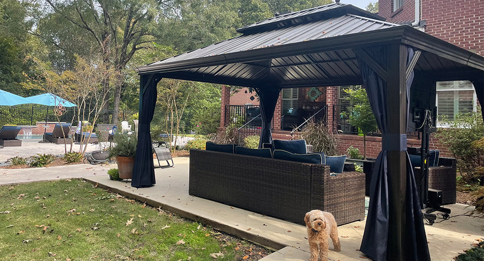 Modern black aluminum gazebo installed on a brick patio, equipped with dark side curtains and wicker outdoor sofa, with a brick house in the background and a beige curly-haired dog in the foreground