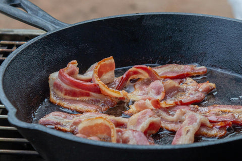 A cast iron pan with bacon strips cooking.
