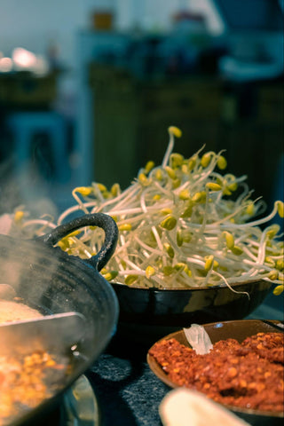 A bowl of appetizing bean sprouts sits next to other ingredients near a pan of cooking food.