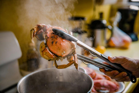A cooked and steaming crab is lifted from a large pot with a pair of tongs.