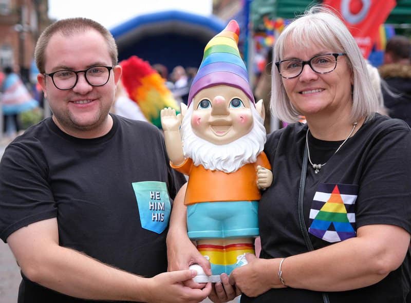 Adam, a white trans man, stands on the left and Louise, a white cis woman, stands on the right. Together they are holding a large pride gnome with a rainbow hat.