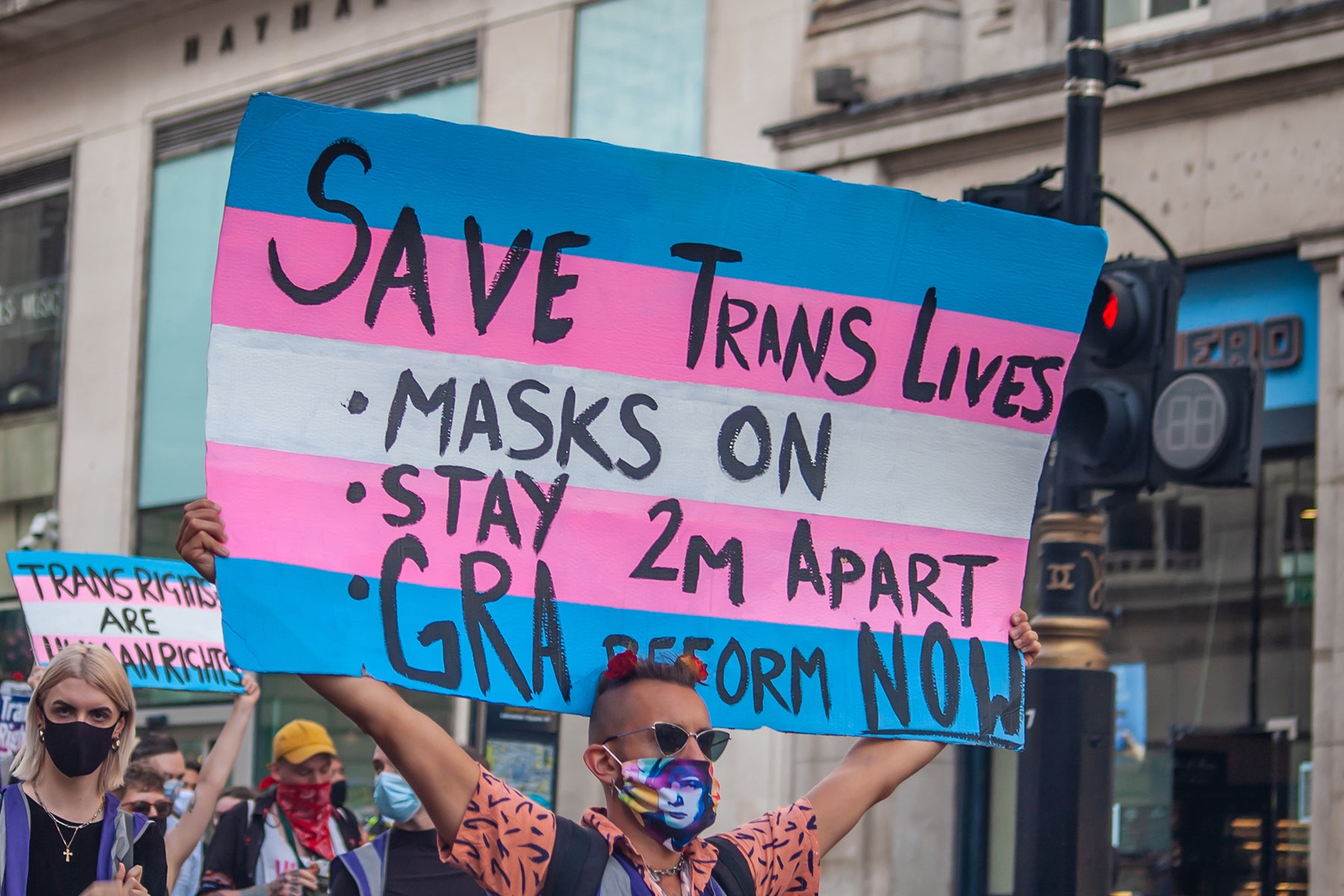 A person holding a protest sign which uses the colours of the transgender flag and reads 'Save Trans Lives - Masks On - Stay 2m Apart - GRA Reform NOW'