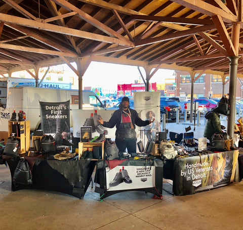 Handmade leather goods displayed on a table including a yellow leather tote, drawstring backpack in black, all made in Detroit by Veterans and Detroiters. There is a Pingree sign and it's at Eastern Market in Detroit. All handmade sustainable leather 