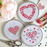 Image of three embroidery hoops with pretty cross stitch heart patterns, threads and scissors