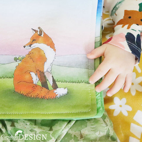 A baby holding a handmade fabric book featuring a fox illustration by Ceridwen Hazelchild.