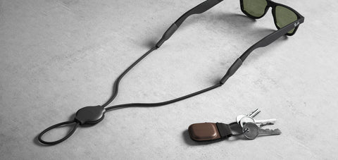 sunglasses attached to Nomad's AirTag glasses holder. next to it is a set of keys with nomad's brown leather airtag holder