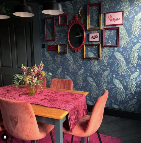 image of dining room with wooden table with pink table runner. Peach tuffted chairs around the table. The wall is covered in a dark wallpaper and decorated with a empty frame gallery wall