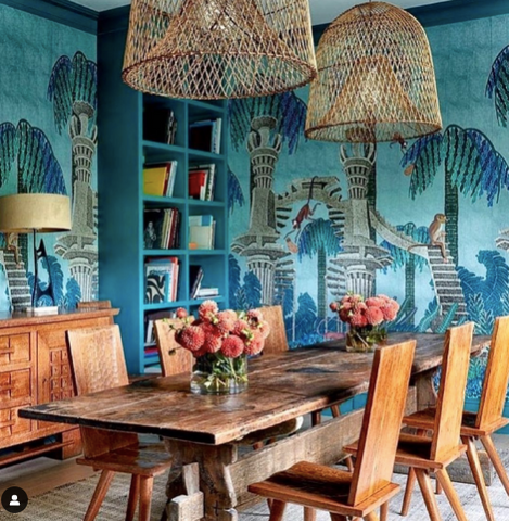 A dining room decorated with a bright blue jungle themed wallpaper, pink flowers and rattan chandeliers