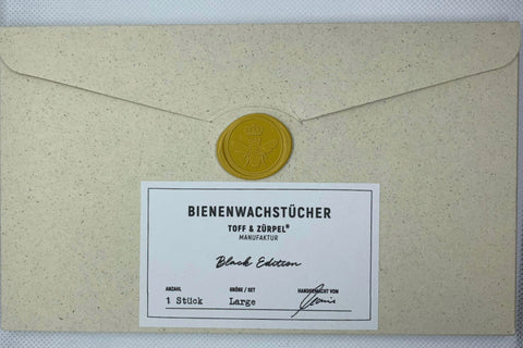 Ein beiger Umschlag mit einem weißen Etikett, auf dem steht: "Bienenwachstücher Toff & Zürpel Manufaktur, Black Edition, 1 Stück, Large". Der Umschlag ist mit einem gelben Wachssiegel verschlossen.