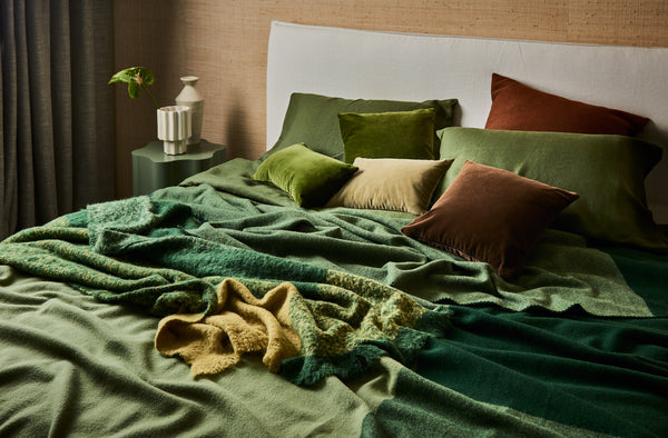 A modern bed dressed with a green merino wool blanket, alpaca throw in green and yellow with assorted pillows. It looks as if someone has just left the frame.