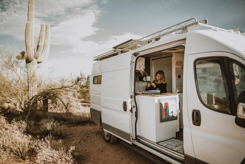 a white campervan is parked in the desert. The side door of the van is open and the owner of Topaz & Pearl is inside making jewelry from her nomadic studio.