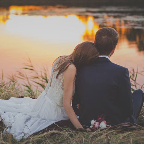 A bride and groom sitting on grass by a river black wedding rings just out of view