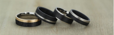 A group of black wedding rings on a table. The black Carbon Fibre rings are inlayed with Palladium, Bronze and Titanium. They are men's wedding bands