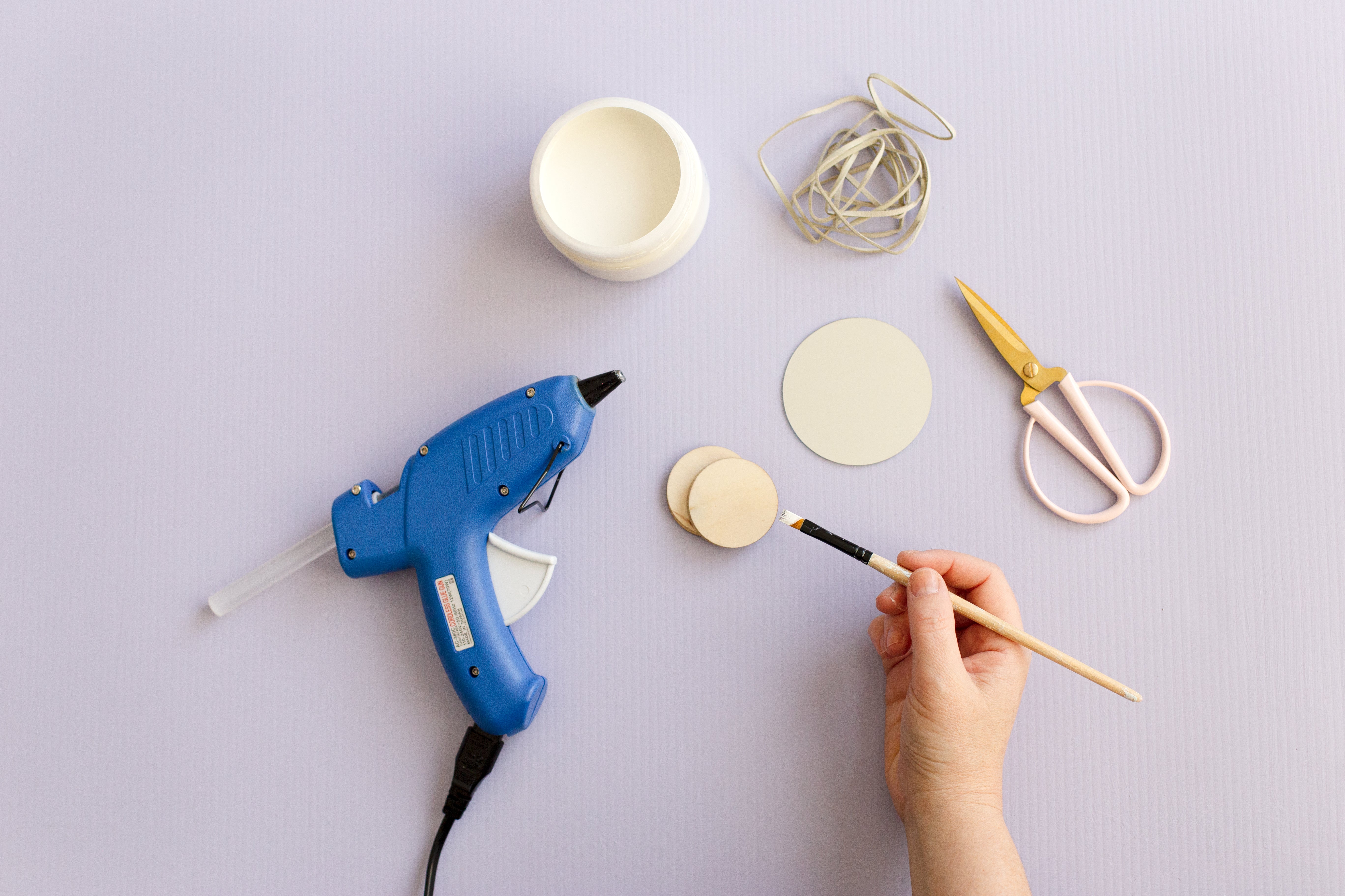 A blue hot glue gun, a pot of white paint, some faux leather string, a pair of pink and gold scissors, a round mirror and some smaller round wooden discs, all flatlayed on a pale lilac background. There's a hand holding a small paintbrush that that has been dipped in the white paint.