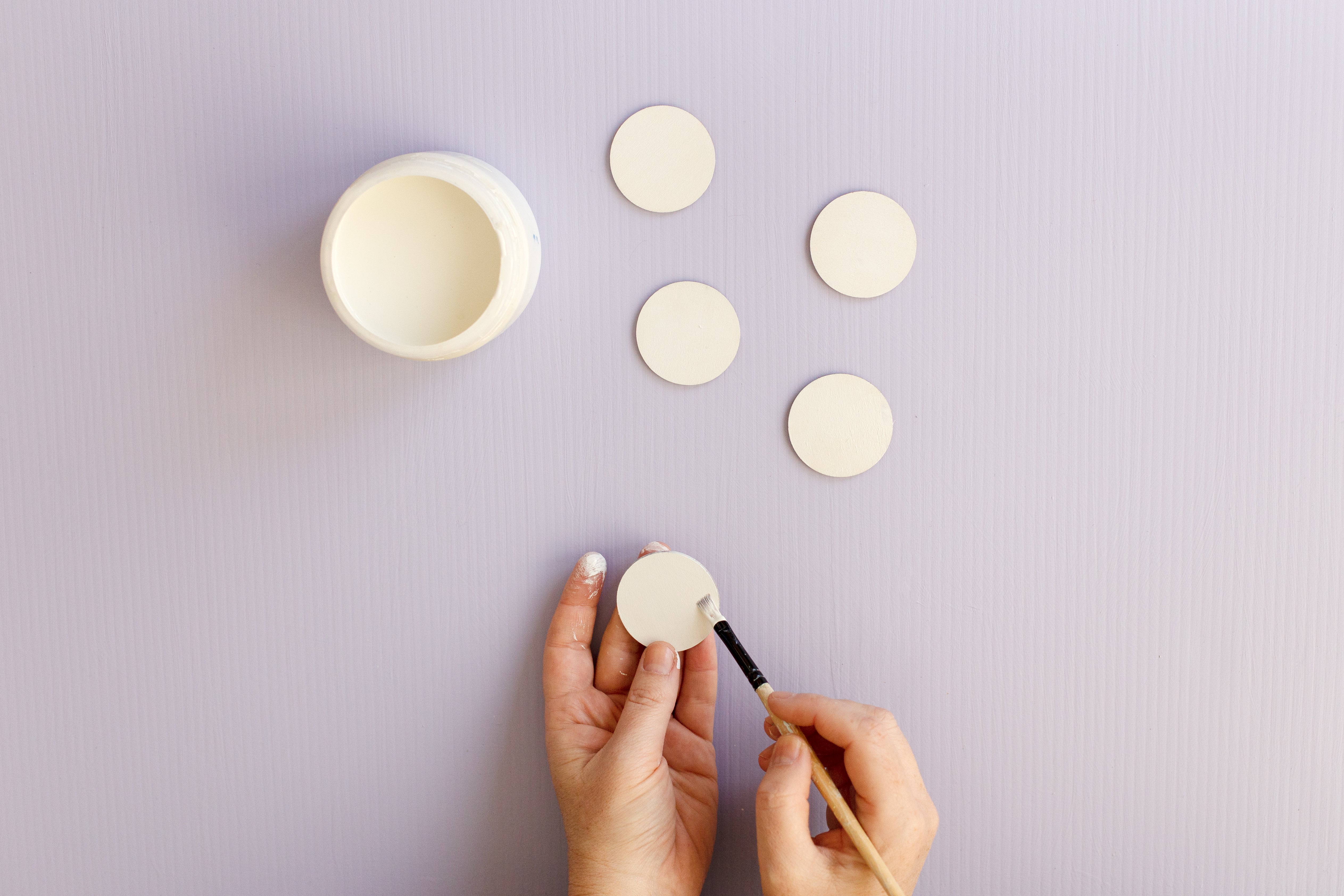 A pot of white paint and four small wooden discs on a purple background. There are two hands holding another small wooden disc, painting them white with a small white paintbrush