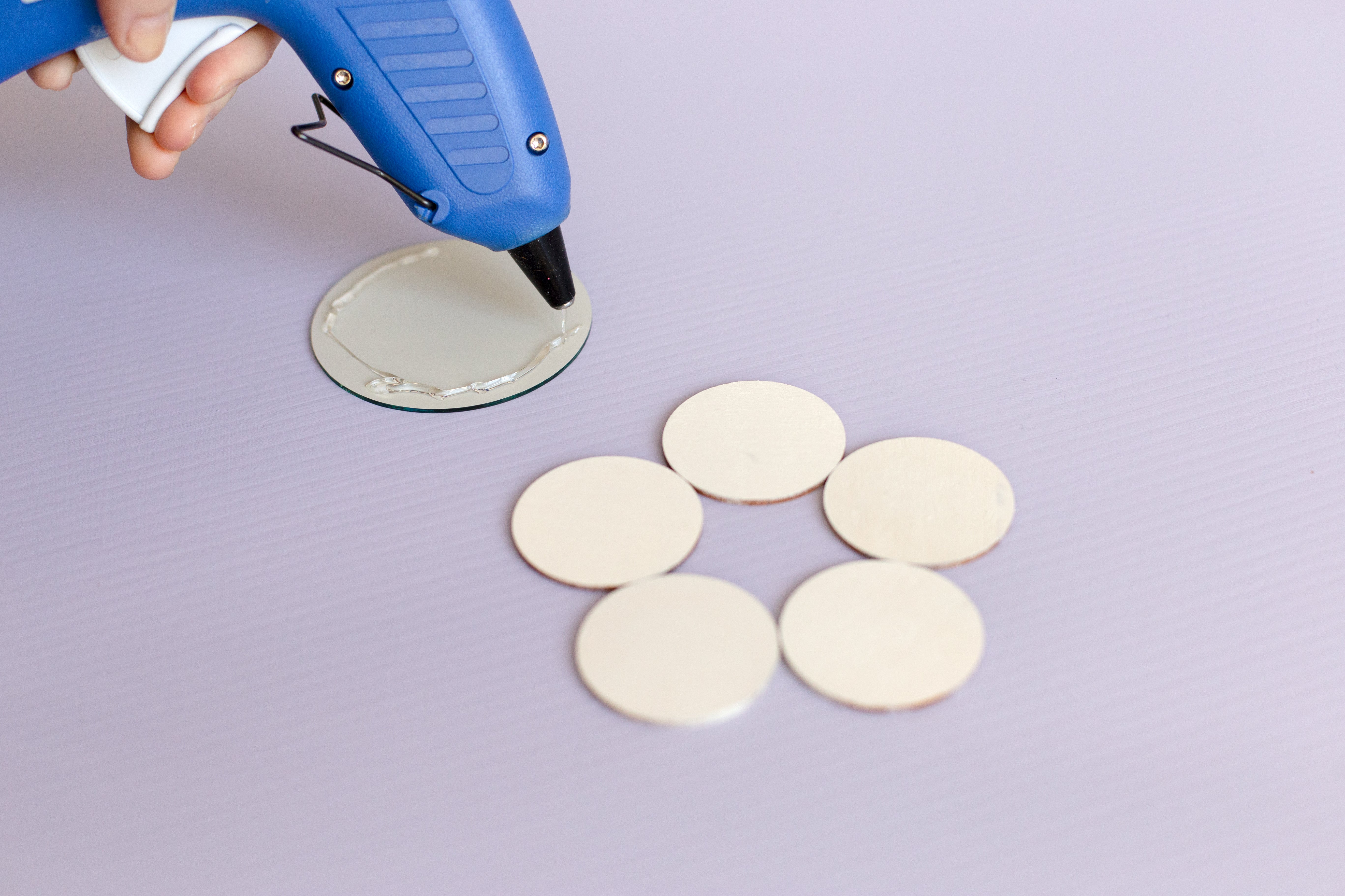 Five small white painted discs are laid out on a pale lilac background. There's a blue hot glue gun in the top left, putting some glue onto the back of a slightly larger, round mirror.