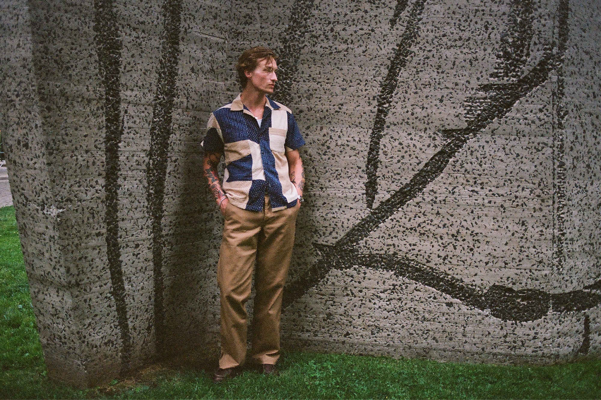 A handsome man in a patchwork shirt stands against a concrete wall.