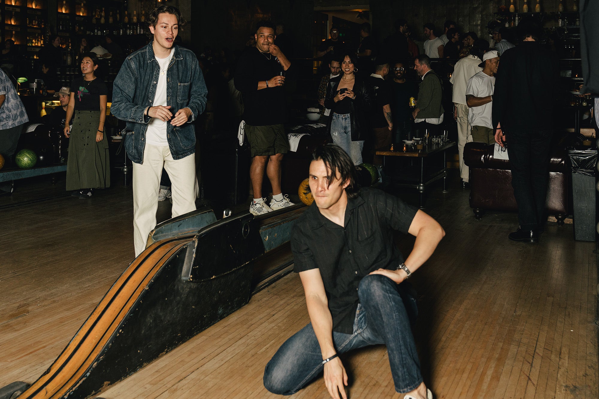 A man crouches on the ground after rolling a bowling ball while others look on.