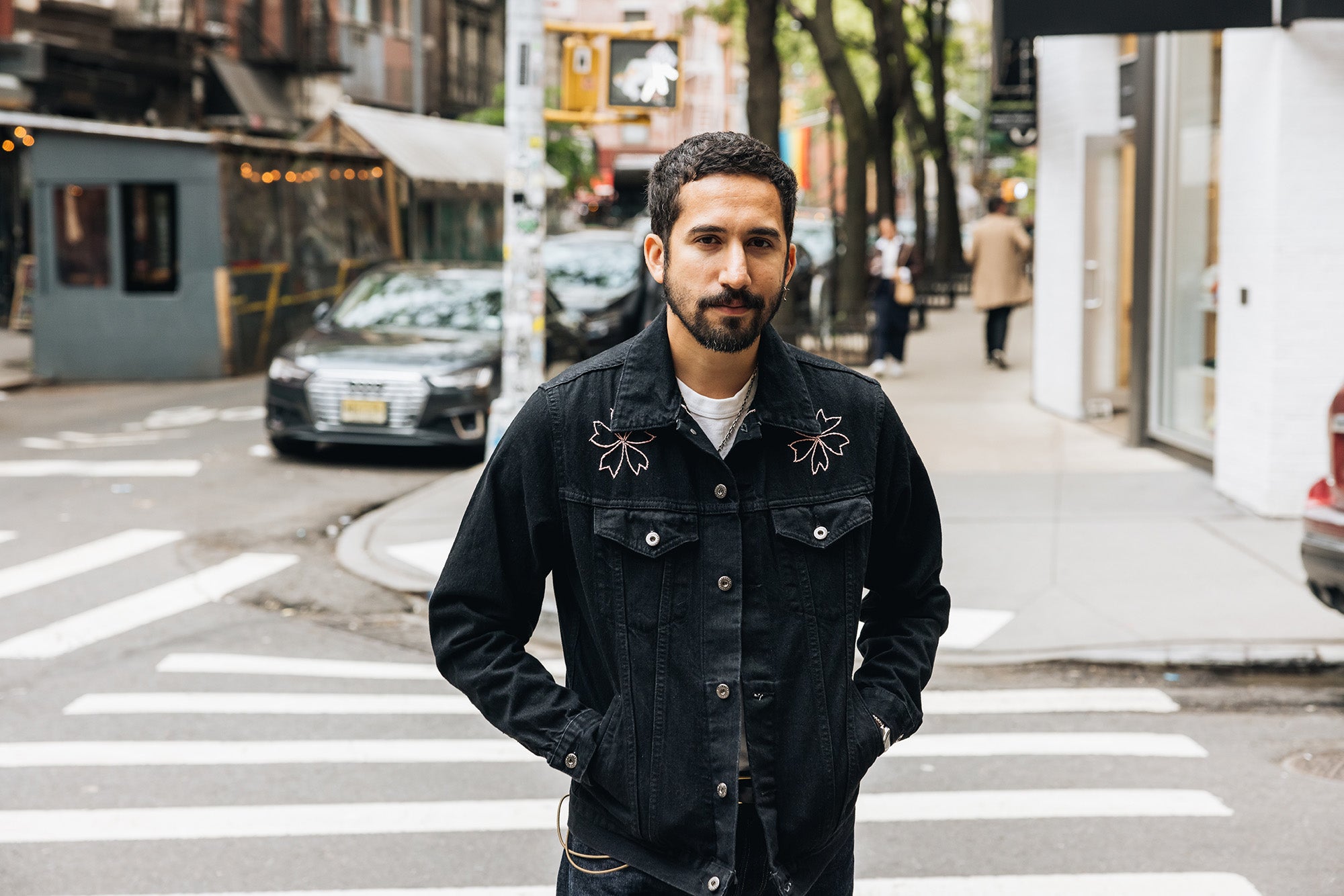 A man in a black denim jacket with sakura blossoms embroidered on the shoulders stands in the street