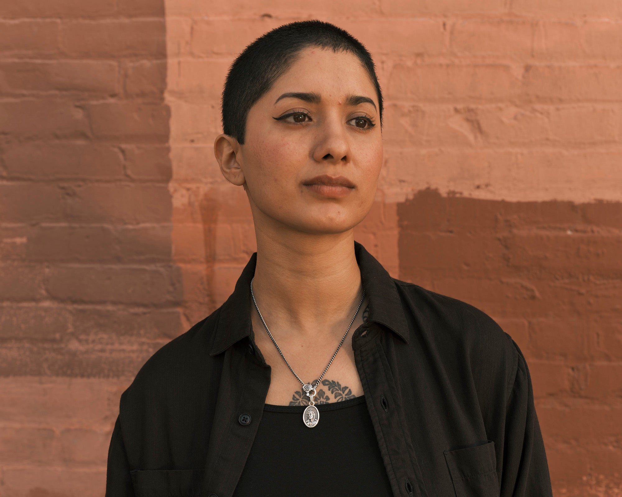 A woman stands in front of a red wall wearing a silver chain with a Jesus pendant on it.