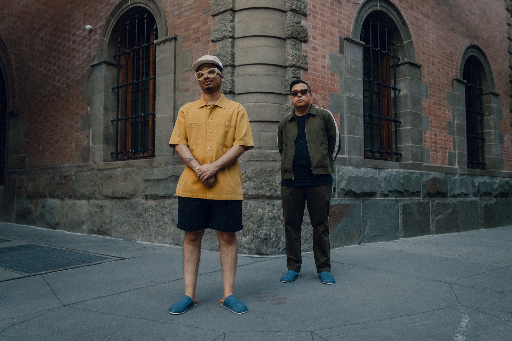 Two men in blue slippers pose on a sidewalk.