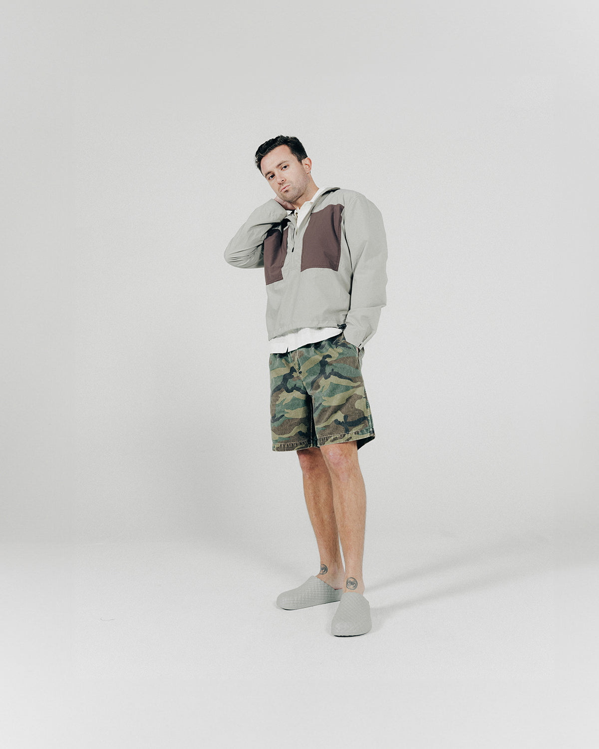 A man poses in a grey nylon pullover shirt and camo shorts.