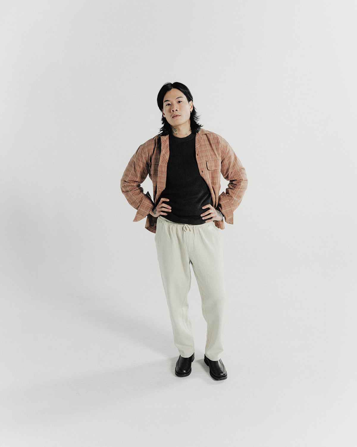 A man poses in an amber button down shirt and cream colored pants.