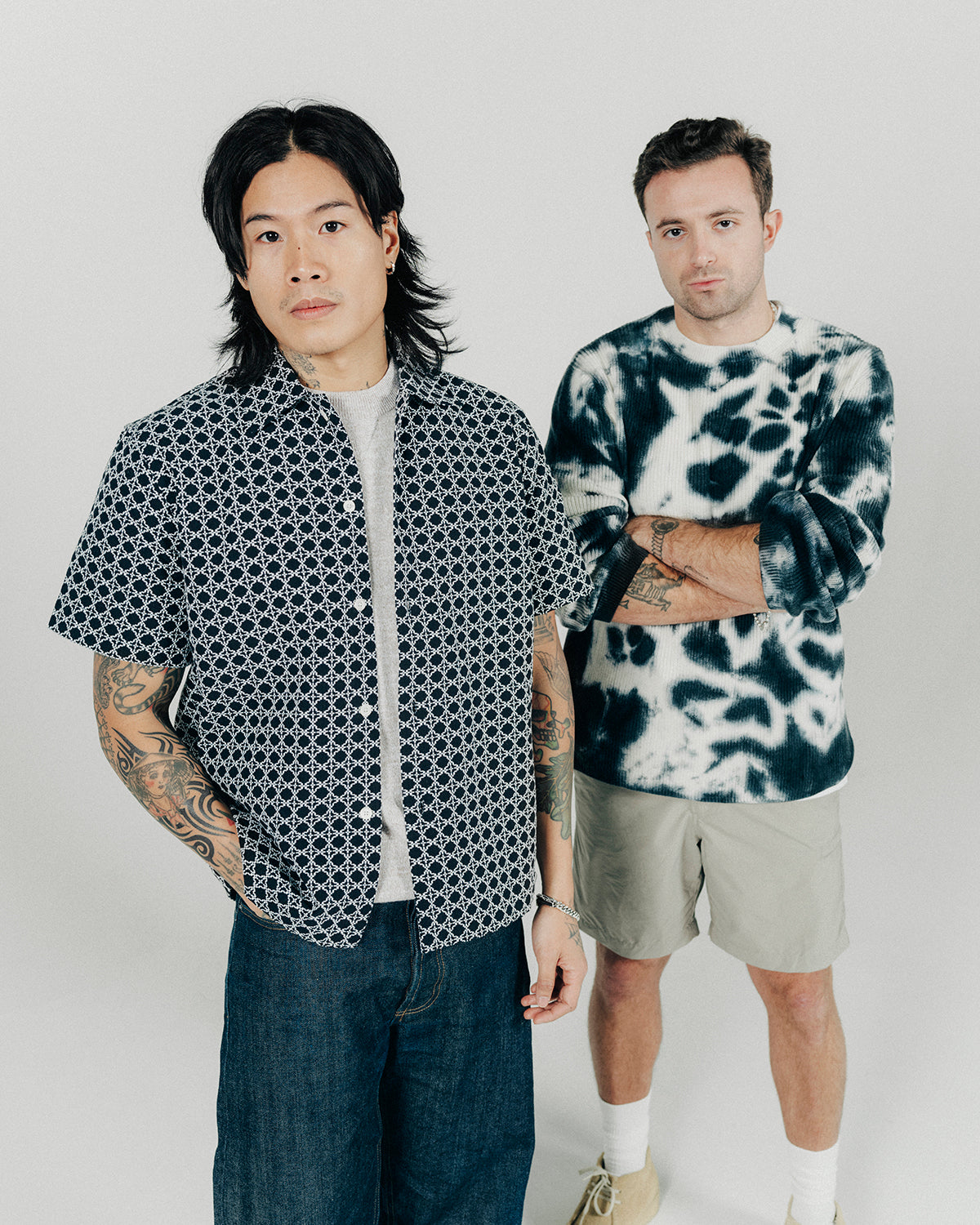 Two men pose, one in a black short sleeve shirt and the other in a tie dyed sweater.