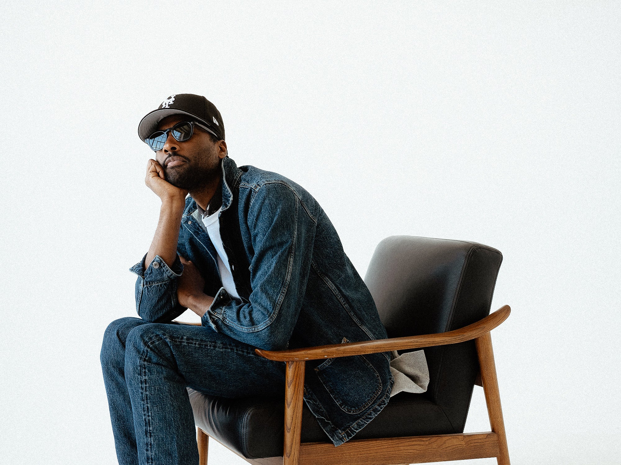 A man in denim jacket and jeans sits on a black leather chair.