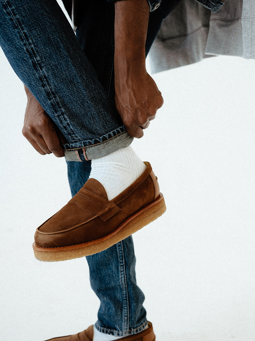 A man adjusts the cuffs on his jeans while showing some brown loafers.