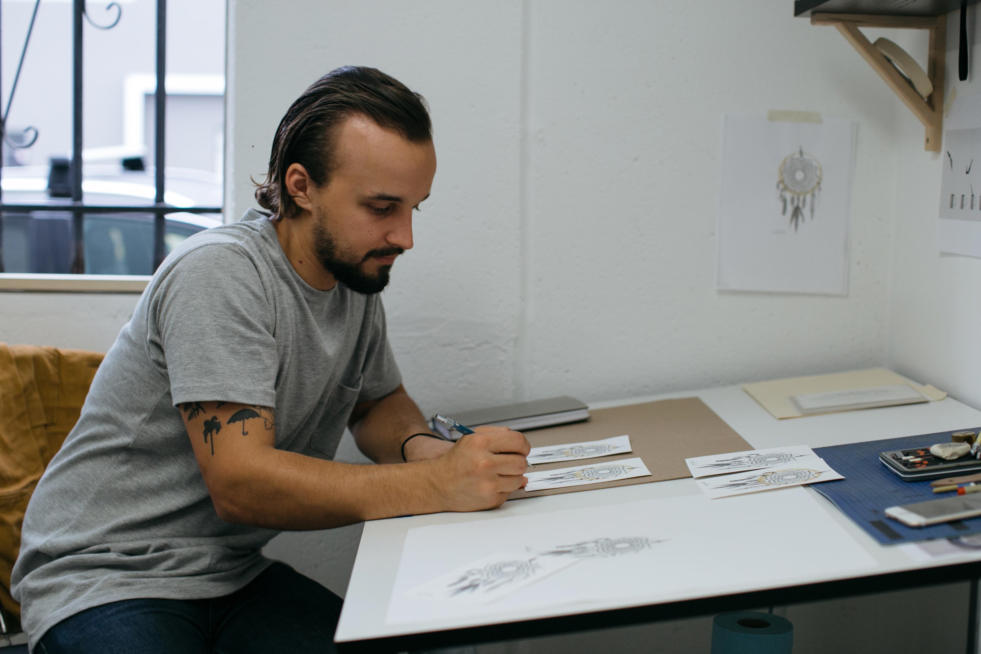 Photo of Carter at work in his studio with a coffee-stain dreamcatcher drawing hanging in the background.