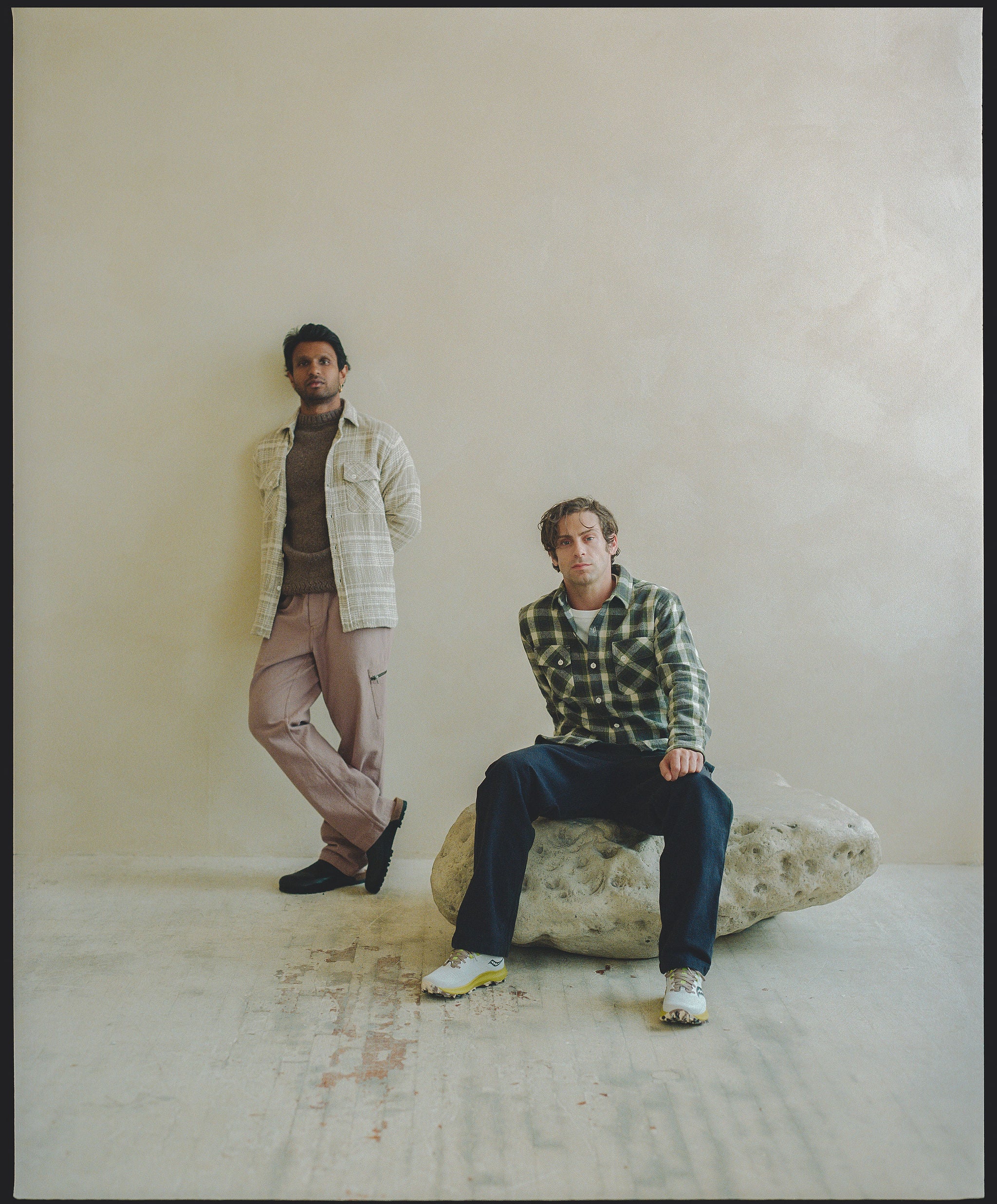 A duo of men in flannel shirts and roomy pants in a neutral colored room.