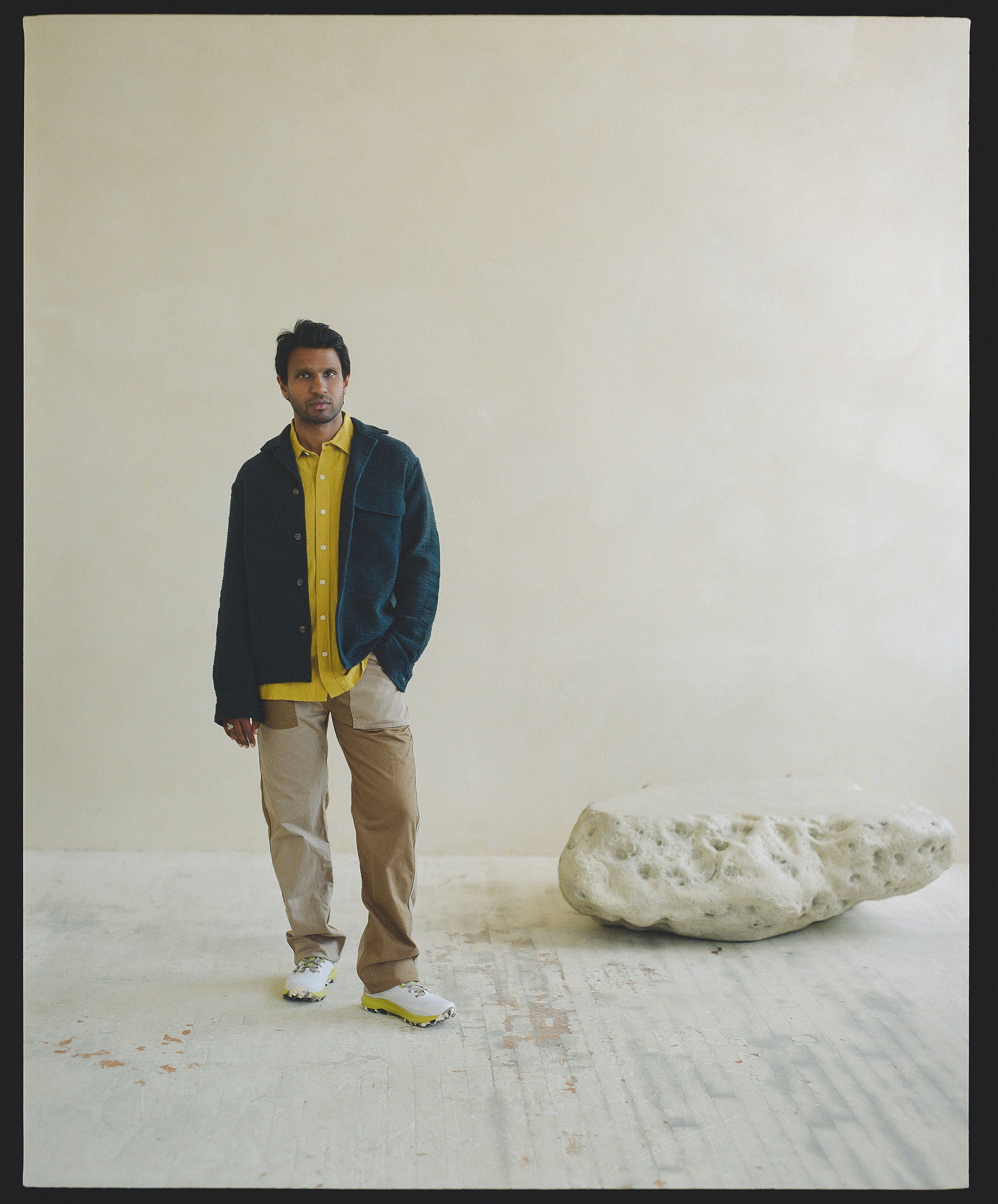 a man in a dark navy overshirt layered over a yellow button down stands next to a rock.