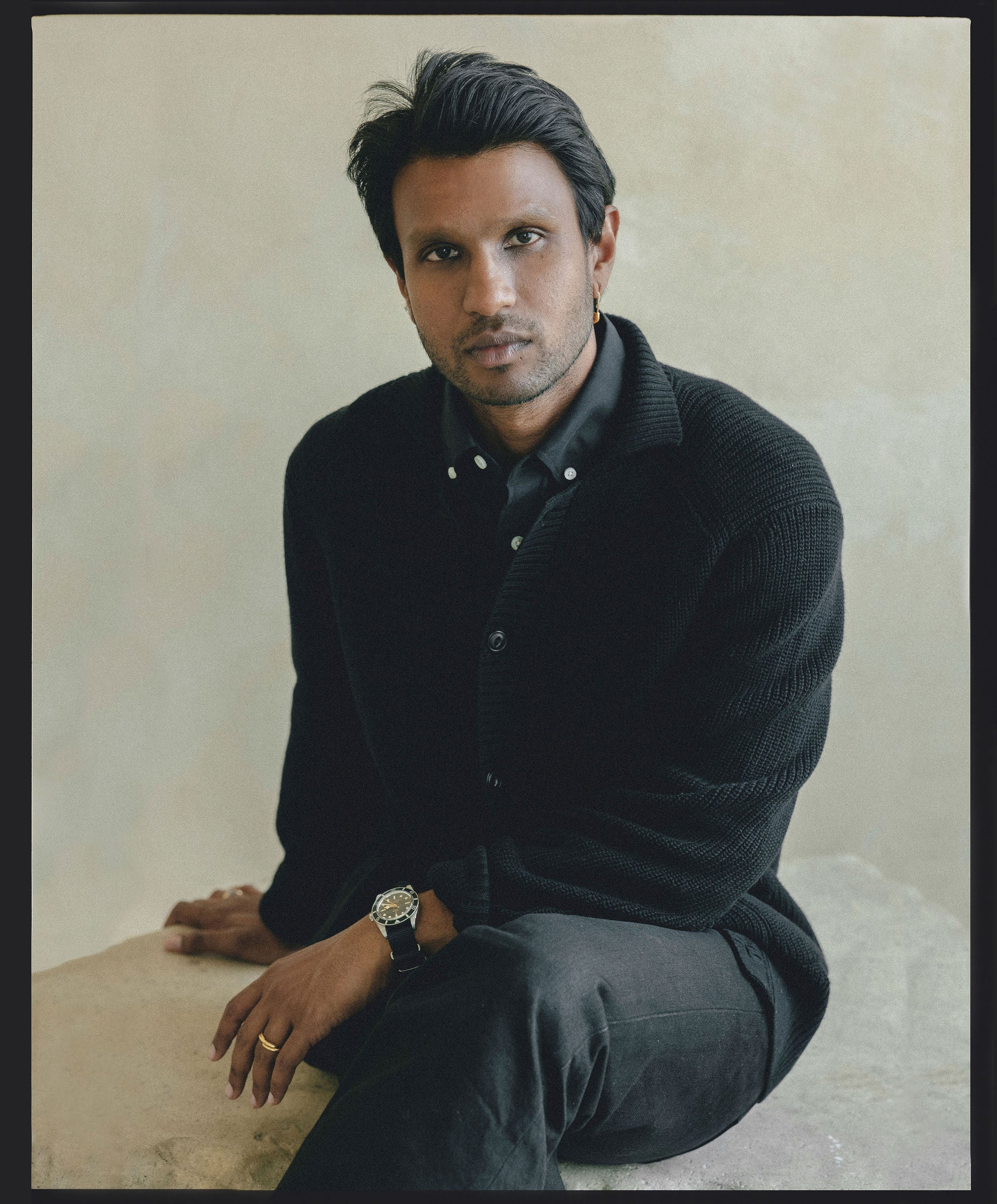 A man in a black shirt and black sweater sits casually on a rock.