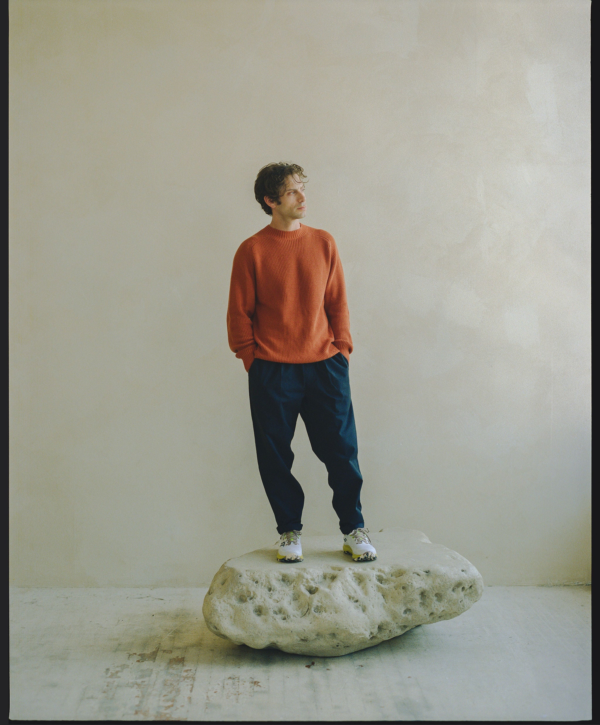 A man in an orange crewneck sweater and navy slouchy pants stands on a rock.