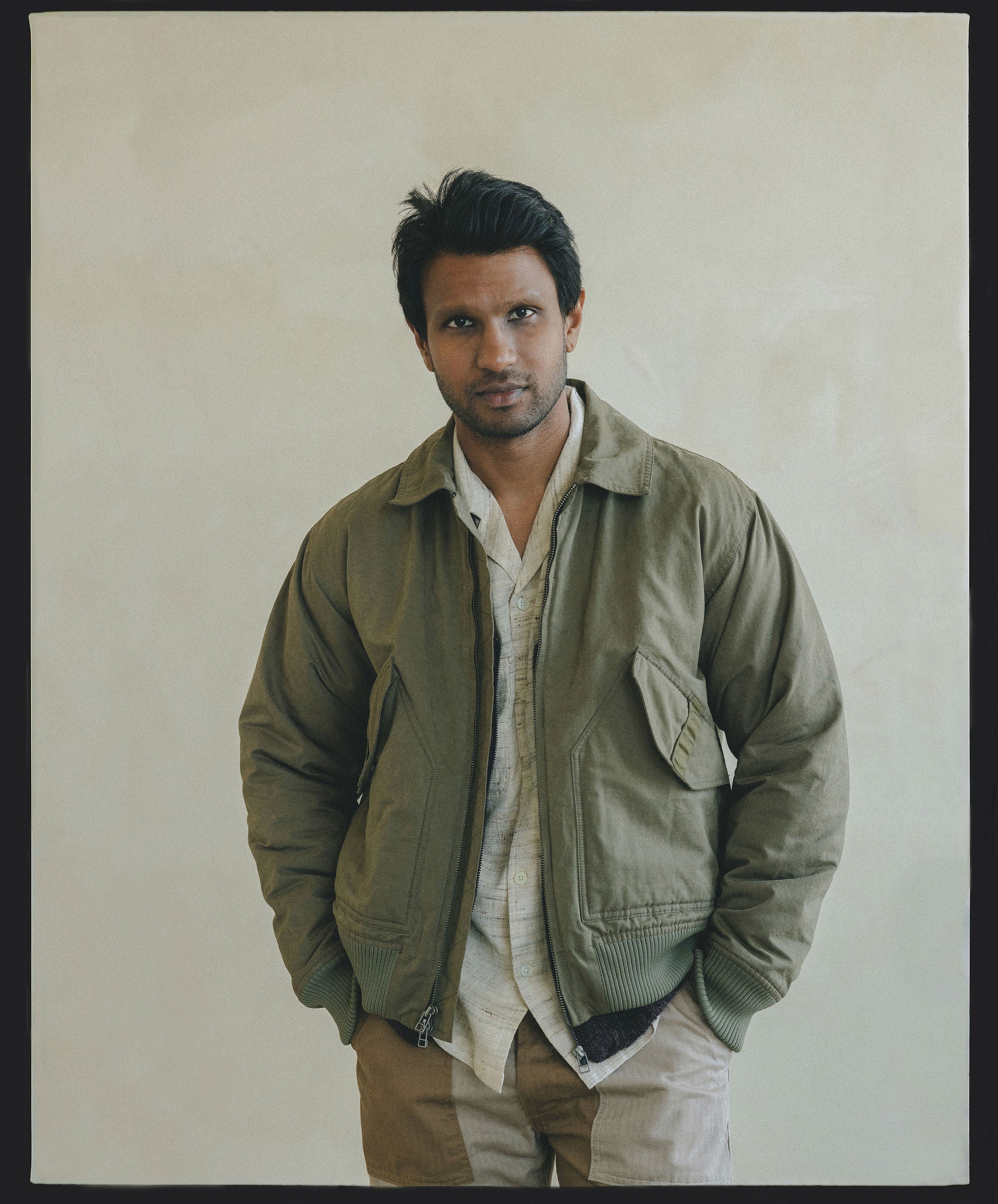 A man in an olive bomber jacket over a cream shirt looks into the camera.